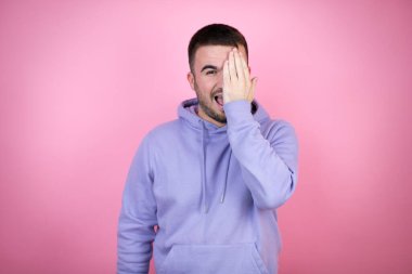 Genç yakışıklı bir adam, pembe bir arka plan üzerine günlük eşofman giyip bir gözünü elinle kapatıyor, yüzünde kendine güvenen bir gülümseme ve sürpriz bir duygu oluşuyor..