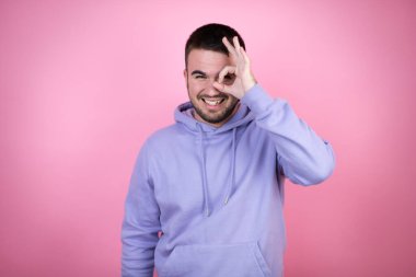 Soyutlanmış pembe arka planda rahat bir eşofman giyen genç yakışıklı adam gülümseyen yüzüyle, gözleriyle parmaklarından bakarak şok oluyor.