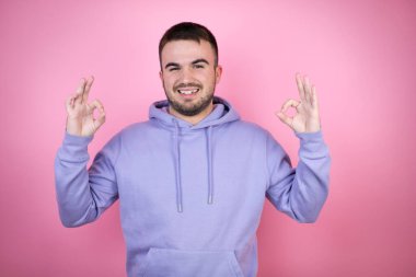İzole edilmiş pembe arka plan üzerine sıradan bir kazak giyen genç yakışıklı adam tamam işareti yapıyor parmaklarıyla ve gülümsüyor, mükemmel bir sembol.