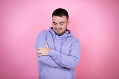 Soyutlanmış pembe arka plan üzerine sıradan bir eşofman giyen genç yakışıklı adam kendine sarılıyor mutlu ve pozitif, kendine güvenen.