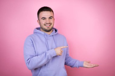İzole edilmiş pembe arka planda sıradan bir eşofman üstü giyen genç yakışıklı bir adam elindeki bir şeyi gösterip işaret ediyor.