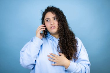İzole edilmiş mavi arka plan üzerine sıradan bir sweatshirt giyen genç ve güzel bir kadın telefonda endişeli bir ifadeyle konuşuyor.