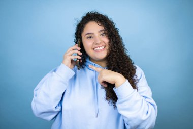 Genç, güzel bir kadın, mavi arka planda rahat bir tişört giyiyor. Gülümsüyor, kulağına taktığı telefonu işaret ediyor.