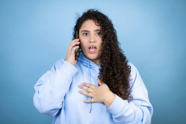İzole edilmiş mavi arka plan üzerine sıradan bir sweatshirt giyen genç ve güzel bir kadın telefonda endişeli bir ifadeyle konuşuyor.