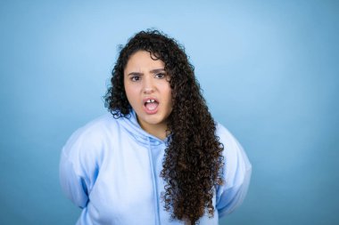 İzole edilmiş mavi arka plan üzerinde sıradan bir eşofman üstü giyen genç ve güzel bir kadın şaşkınlık ve şaşkınlık dolu bir ifade, korku ve heyecanlı bir yüz ifadesiyle şok olmuş ve korkmuştu..