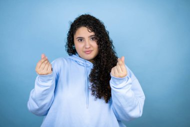 İzole edilmiş mavi arka plan üzerine sıradan bir sweatshirt giyen genç güzel bir kadın. Elleriyle para hareketi yapıyor. Maaş ödemesi istiyor.