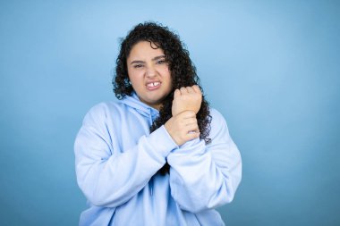 İzole edilmiş mavi arka plan üzerine sıradan eşofman üstü giyen genç ve güzel bir kadın. Ellerinde ve parmaklarında ağrı, eklem iltihabı.