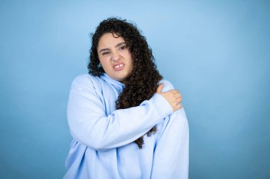 Genç ve güzel bir kadın, izole edilmiş mavi arka planda, omzunda ağrı ve acı dolu bir ifadeyle gündelik eşofman üstü giyiyor.
