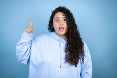 İzole edilmiş mavi arka plan üzerinde sıradan bir eşofman üstü giyen genç ve güzel bir kadın parmağını kafasında gezdirmiş ve bir fikri olduğunu düşünüyor..