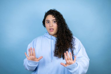 İzole edilmiş mavi arka plan üzerinde sıradan bir eşofman üstü giyen genç ve güzel bir kadın korku ifadesi karşısında korkmuş ve dehşete düşmüş. El hareketlerini durdur, şok içinde bağır. Panik konsepti.