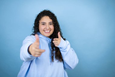İzole edilmiş mavi arka plan üzerine günlük eşofman giyen genç ve güzel bir kadın sana ve kameraya parmaklarıyla bakıyor, mutlu ve mutlu gülümsüyor.