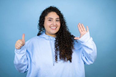 İzole edilmiş mavi arka plan üzerine sıradan bir kazak giyen genç ve güzel bir kadın altı numaralı parmaklarıyla gülümseyerek kendinden emin ve mutlu bir şekilde onu işaret ediyor.