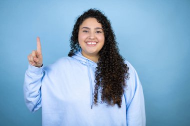 İzole edilmiş mavi arka plan üzerine sıradan bir sweatshirt giyen genç ve güzel bir kadın bir numaralı parmaklarını gösterirken kendinden emin ve mutlu bir şekilde gülümsüyor.