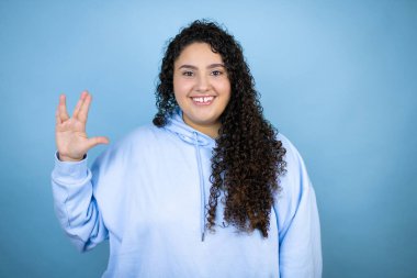 İzole edilmiş mavi arka plan üzerine sıradan bir kazak giyen genç ve güzel bir kadın Star Trek ucube sembolü yapıyor.