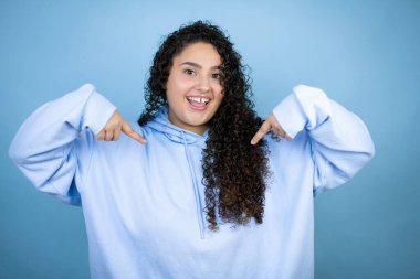 İzole edilmiş mavi arka plan üzerinde sıradan bir eşofman üstü giyen genç ve güzel bir kadın. Kendine güvenen, yüzünde bir gülümseme, kendini gururla ve mutlu bir şekilde gösteren bir kadın..