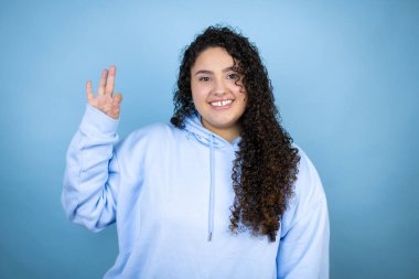 İzole edilmiş mavi arka plan üzerine sıradan bir kazak giyen genç ve güzel bir kadın tamam işareti yapıyor parmaklarıyla ve gülümseyerek, mükemmel bir sembol.