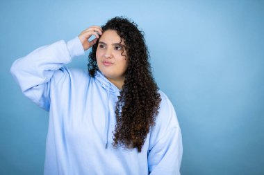 İzole edilmiş mavi arka plan kafa karışıklığı ve soru hakkında merak üzerine sıradan bir sweatshirt giyen genç ve güzel bir kadın. Şüpheyle karışık, kafa kafaya düşünerek.
