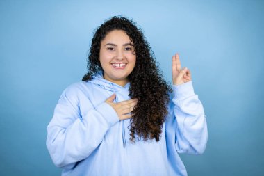 Genç, güzel bir kadın, izole edilmiş mavi arka plan üzerine sıradan bir eşofman üstü giyiyor. Gülümsüyor, elleri göğsünde ve parmakları havada küfrediyor, sadakat yemini ediyor.