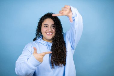 Genç, güzel bir kadın, mavi arka planda rahat bir kazak giyip gülümsüyor ve elleri ve parmakları mutlu yüzlü bir çerçeve yapıyor.