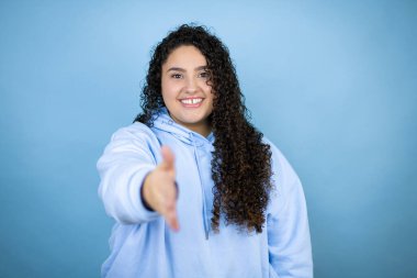 İzole edilmiş mavi arka plan üzerinde sıradan bir eşofman üstü giyen genç ve güzel bir kadın selamlaşma ve dostça bir şekilde el sıkışma sunuyor.