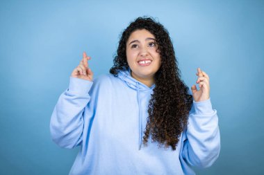 İzole edilmiş mavi arka plan üzerine sıradan bir kazak giyen genç ve güzel bir kadın umut dolu gülümseyen ve yan bakan bir parmak işareti yapıyor.