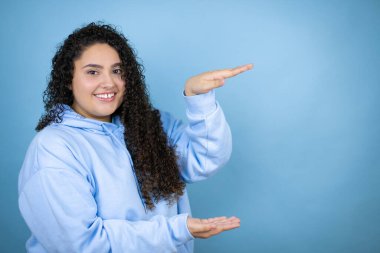 İzole edilmiş mavi arka plan jestlerinin üzerine gündelik eşofman giyen genç ve güzel bir kadın. Ellerinde büyük ve büyük bir işaret, ölçü sembolü var. Gülümsüyor. Ölçüm konsepti.