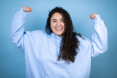 Genç, güzel bir kadın, mavi arka plan üzerine günlük eşofman üstü giyerek mutlu ve heyecanlı bir şekilde kollarını kaldırıp, gülümseyerek ve başarı için çığlık atarak kazanan bir jest yapıyor..