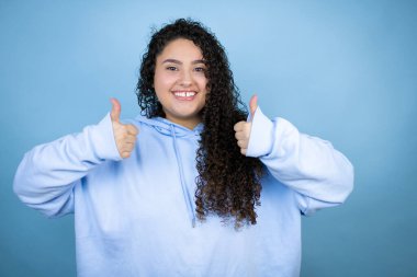 Genç, güzel bir kadın, mavi arka planda günlük eşofman üstü giyerek gülümsüyor ve baş parmaklarıyla işaret veriyor.