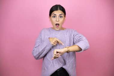 Güzel bir kadın pembe arka planda sıradan bir mor kazak giyiyor. Geç olduğu için şaşırıp saatini işaret ediyor.