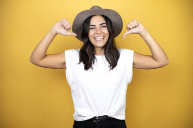 Beautiful woman wearing casual white t-shirt and a hat standing over yellow background looking confident with smile on face, pointing oneself with fingers proud and happy.