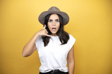 Güzel bir kadın, günlük beyaz tişört ve sarı arka planda duran bir şapka giyer telefon hareketlerini elle ve parmaklarla yapar telefonda konuşur gibi yapar.