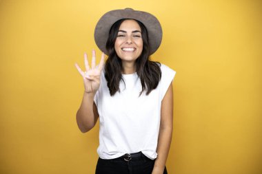 Güzel bir kadın, günlük beyaz tişört ve sarı arka planda duran bir şapka giyer ve dört numarayı parmaklarıyla gösterirken kendinden emin ve mutlu bir şekilde gülümser.