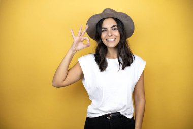 Beautiful woman wearing casual white t-shirt and a hat standing over yellow background doing ok sign with fingers and smiling, excellent symbol