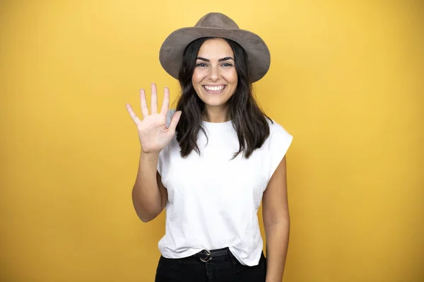 漂亮的女人穿着随意的白色T恤 头戴一顶帽子 站在黄色的背景上 带着自信和快乐的笑容 用手指着五号 — 图库照片