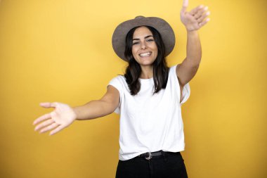 Güzel bir kadın, günlük beyaz tişört ve sarı arka planda duran bir şapka giyer ve kameraya bakar. Sarılmak için kollarını açar ve gülümser.