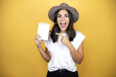 Güzel bir kadın, günlük beyaz tişört ve sarı arka planda duran bir şapka giyiyor. Gülümsüyor ve elinde boş bir defter gösteriyor.