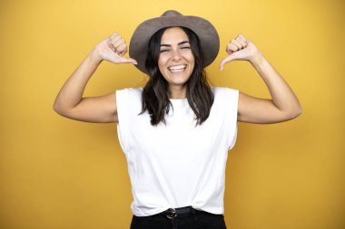 Beautiful woman wearing casual white t-shirt and a hat standing over yellow background looking confident with smile on face, pointing oneself with fingers proud and happy.