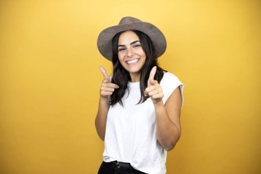 Beautiful woman wearing casual white t-shirt and a hat standing over yellow background pointing to you and the camera with fingers, smiling positive and cheerful