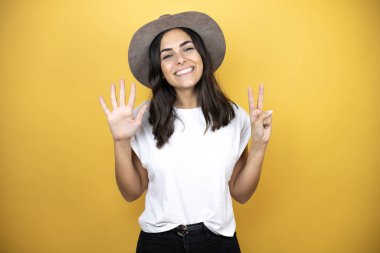 Güzel bir kadın, günlük beyaz tişört ve sarı arka planda duran bir şapka giyer ve yedinci parmaklarıyla işaret ederken kendinden emin ve mutlu bir şekilde gülümser.