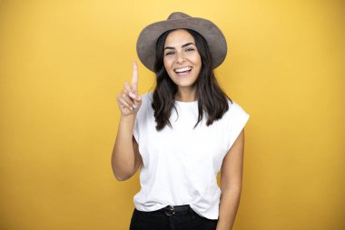 Güzel bir kadın, günlük beyaz tişört ve sarı arka planda duran bir şapka giyer ve parmaklarıyla bir numarayı gösterirken kendinden emin ve mutlu bir şekilde gülümserdi.