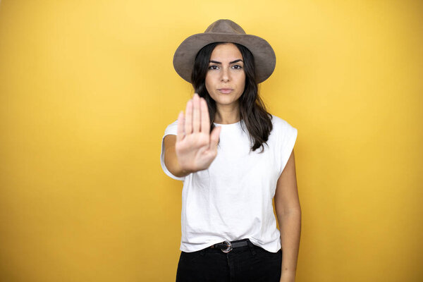 Beautiful woman wearing casual white t-shirt and a hat standing over yellow background serious and doing stop sing with palm of the hand.