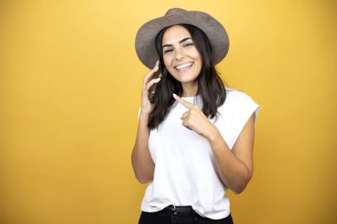 Güzel bir kadın, günlük beyaz tişört ve sarı arka planda duran bir şapka giyer. Gülümser, kulağına taktığı telefonu işaret eder.