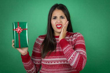 Genç, güzel bir kız, diş ağrısı ya da diş hastalığından dolayı izole edilmiş beyaz bir arkaplanda hediyesini tutuyor acı dolu bir ifadeyle ağıza dokunuyor.