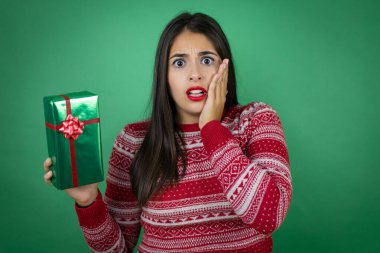 Genç ve güzel bir kız hediyesini izole edilmiş beyaz arka planda tutuyor. Çığlık atıyor ve elleri ağzında şaşırıyor.