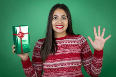 Genç, güzel bir kız hediyesini izole edilmiş beyaz arka planda tutarken ve parmaklarıyla beş numarayı gösterirken kendinden emin ve mutlu gülümserken.