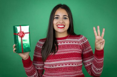 Genç, güzel bir kız, kendine güvenen ve mutlu gülümserken beyaz arka planda hediyesini tutuyor ve üç numarayı işaret ediyor.