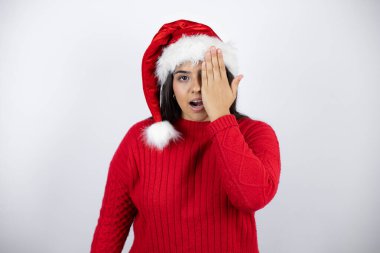 Beyaz arka planda Noel Baba şapkası takan genç güzel bir kadın bir gözünü eliyle kapatıyor, yüzünde kendine güvenen bir gülümseme ve sürpriz bir duygu var..