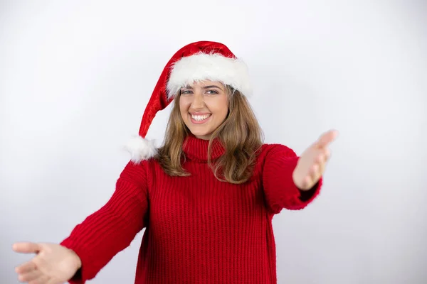年轻漂亮的金发女人 身穿红色休闲衫 头戴白色背景的圣诞帽 看着镜头 张开双臂拥抱对方 — 图库照片