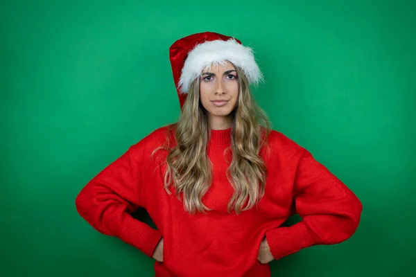 年轻美丽的女孩戴着圣诞圣诞圣诞礼帽 带着礼物 而不是孤零零的绿色背景 怀疑和紧张 脸上带着不赞成的表情 胳膊插在腰部 — 图库照片