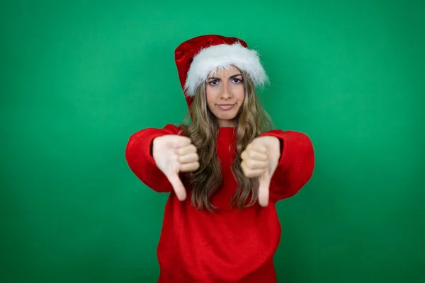 年轻美丽的女孩戴着圣诞圣诞礼帽 带着礼物披着孤零零的绿色背景 满脸怒容 负面的迹象表明她不喜欢小拇指朝下 — 图库照片